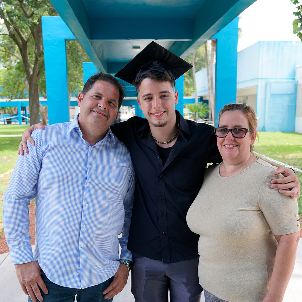 Miami-Dade Acceleration Academies graduate stands wearing cap and gown with his parents.